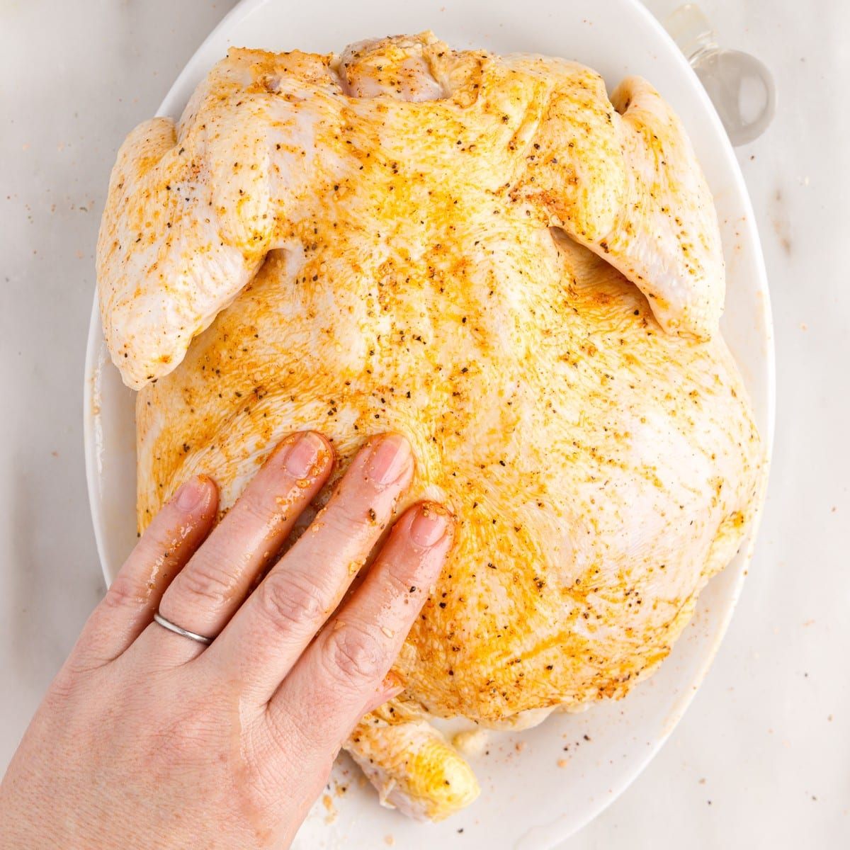 Raw whole chicken being rubbed with olive oil and seasonings by hand on a white platter while preparing Air Fryer Whole Chicken.