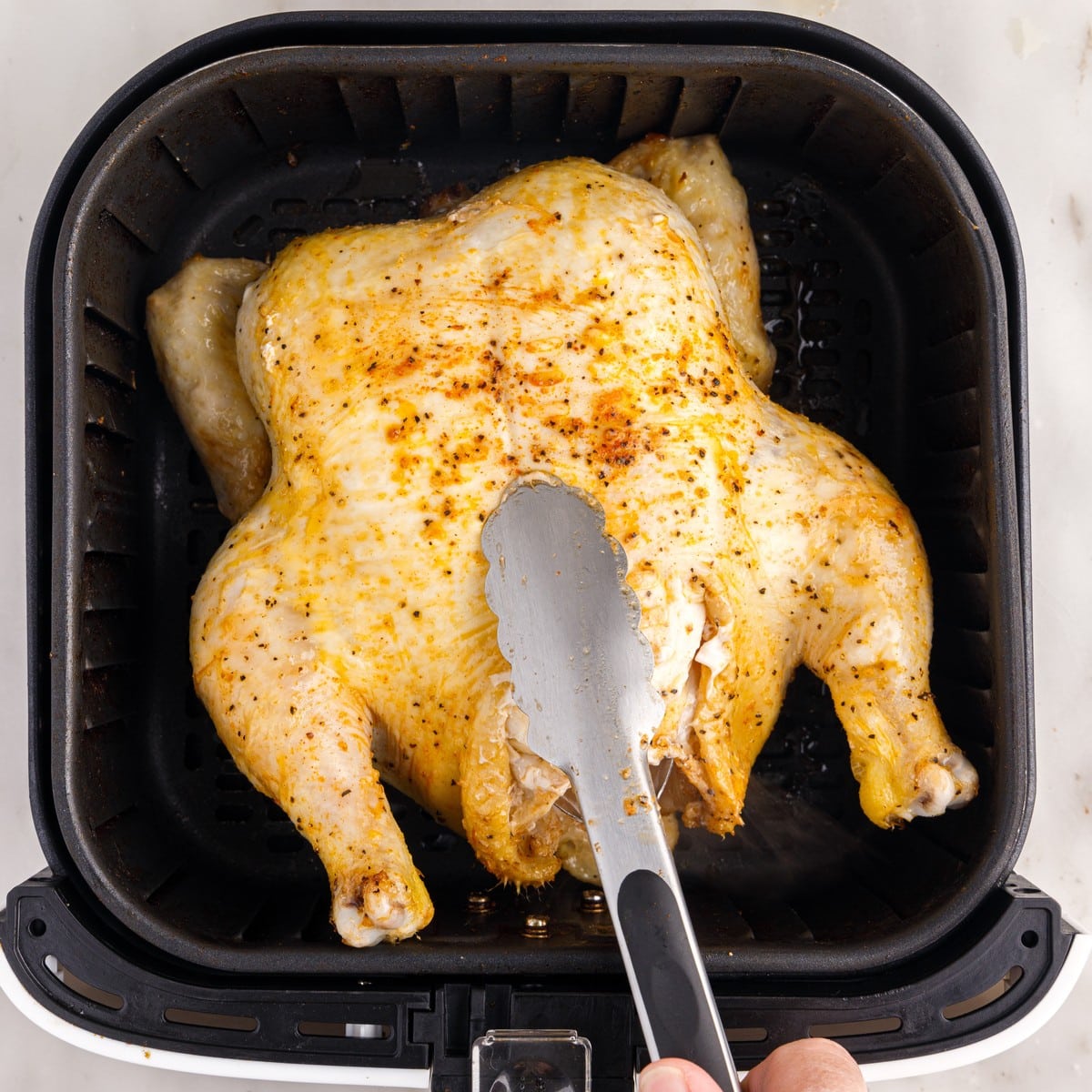 Seasoned raw whole chicken being placed breast-side down into a black air fryer basket with kitchen tongs before cooking.