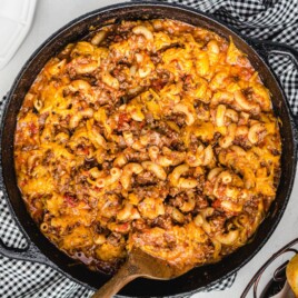 Overhead view of American Chop Suey in a black cast-iron skillet with ground beef, elbow macaroni, tomato sauce, and melted cheddar cheese.