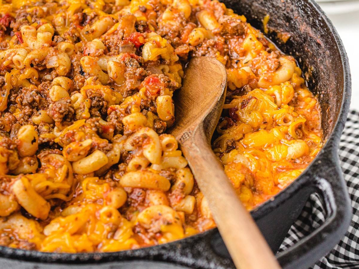 Close-up of cheesy American Chop Suey with elbow macaroni, ground beef, tomatoes, and melted cheddar cheese in a cast-iron skillet with a wooden spoon.