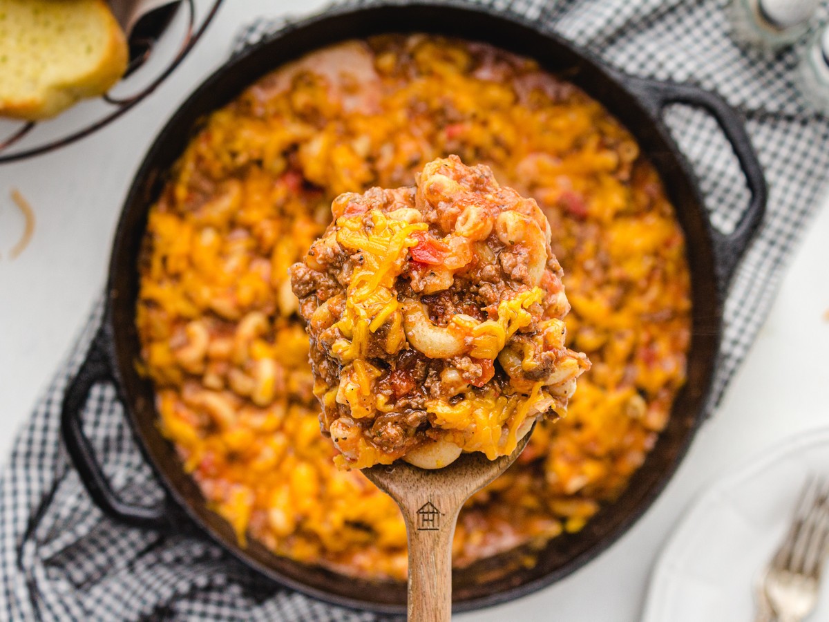 Wooden spoon lifting a hearty serving of American Chop Suey with ground beef, elbow macaroni, tomato sauce, and melted cheddar cheese from a cast-iron skillet.