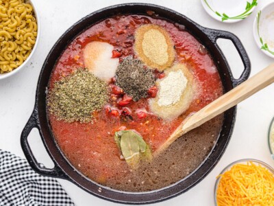 Overhead view of American Chop Suey sauce ingredients in a cast-iron skillet, including tomato sauce, diced tomatoes, Italian seasoning, black pepper, garlic powder, onion powder, salt, bay leaves, and liquid before simmering.