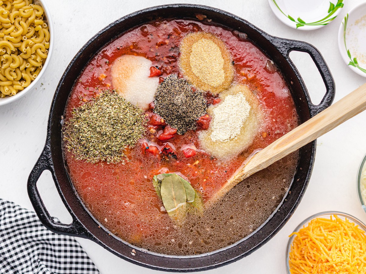 Overhead view of American Chop Suey sauce ingredients in a cast-iron skillet, including tomato sauce, diced tomatoes, Italian seasoning, black pepper, garlic powder, onion powder, salt, bay leaves, and liquid before simmering.