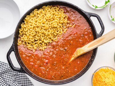 Uncooked elbow macaroni added to the seasoned tomato and ground beef mixture in a cast-iron skillet while preparing American Chop Suey.