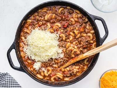 American Chop Suey simmering in a cast-iron skillet with ground beef, elbow macaroni, tomatoes, and a mound of shredded cheese being added before melting.