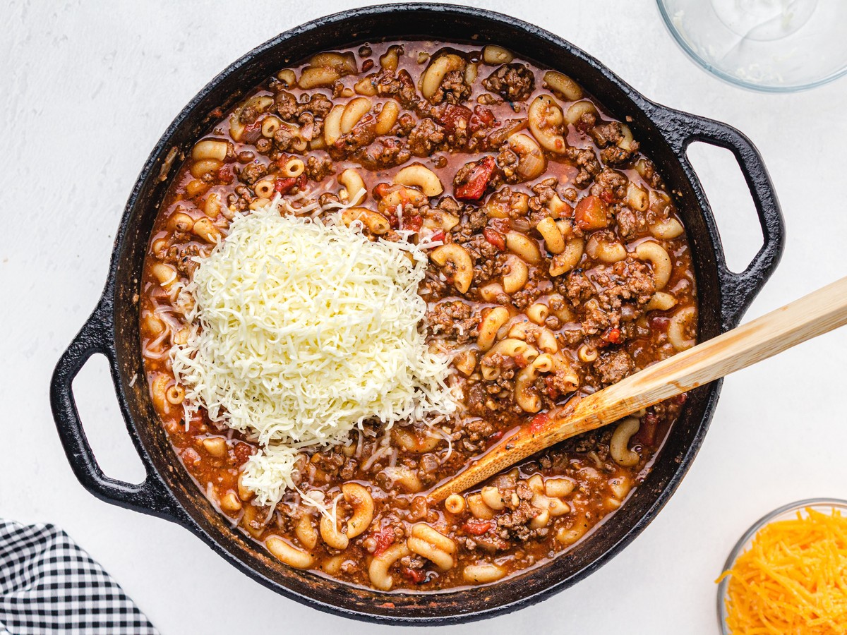 American Chop Suey simmering in a cast-iron skillet with ground beef, elbow macaroni, tomatoes, and a mound of shredded cheese being added before melting.