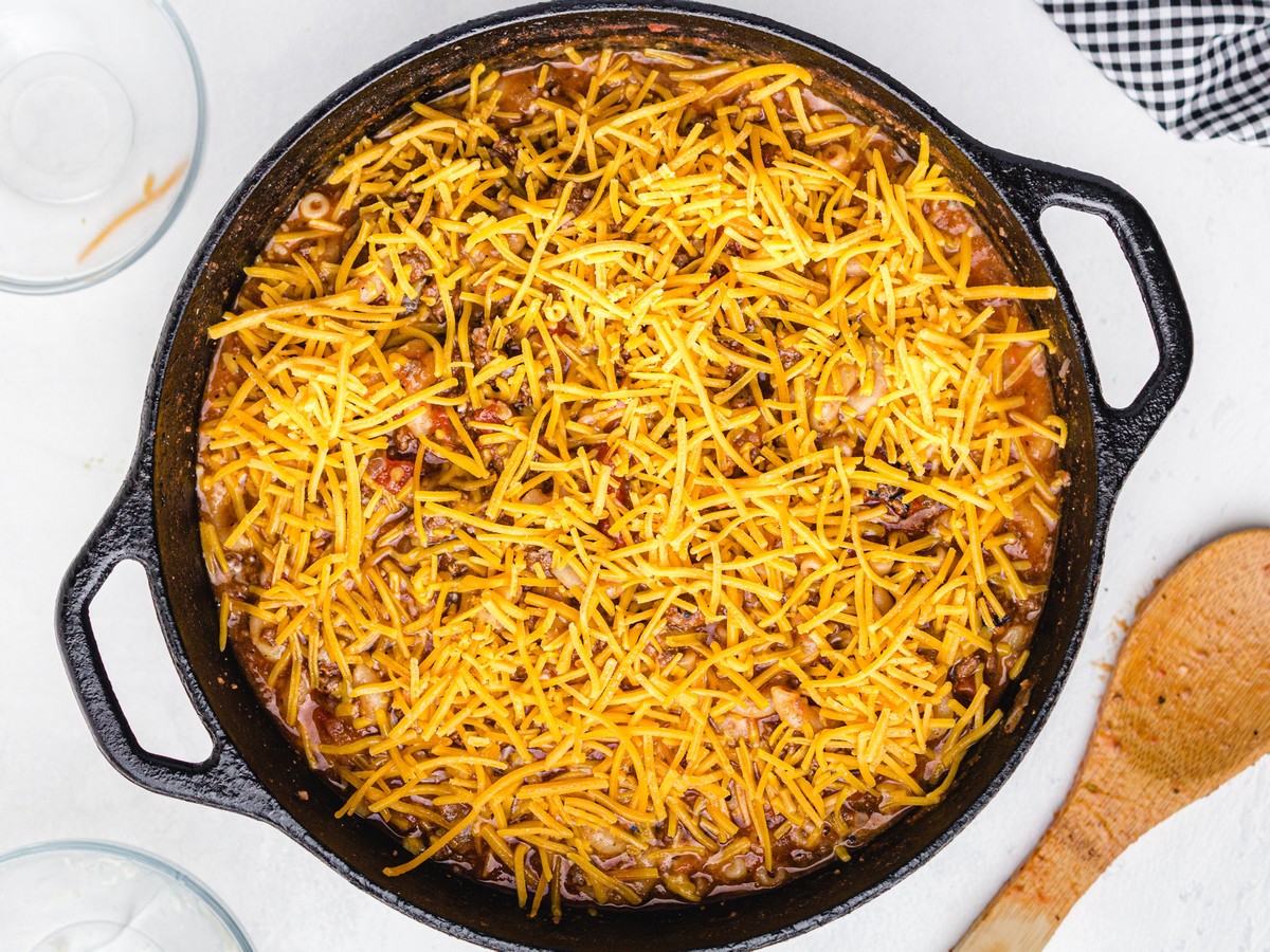 Overhead view of American Chop Suey in a cast-iron skillet topped with an even layer of shredded cheddar cheese before melting.