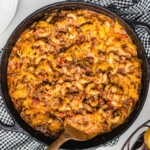 Finished American Chop Suey in a cast-iron skillet with elbow macaroni, ground beef, tomato sauce, and melted cheddar cheese, served with garlic bread.