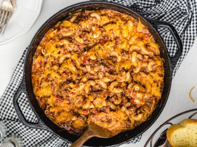 Finished American Chop Suey in a cast-iron skillet with elbow macaroni, ground beef, tomato sauce, and melted cheddar cheese, served with garlic bread.