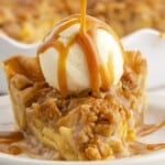 Slice of Caramel Apple Pie topped with a scoop of vanilla ice cream while warm caramel sauce is poured over the cinnamon-spiced apple filling and crumb topping.