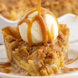 Slice of Caramel Apple Pie topped with a scoop of vanilla ice cream while warm caramel sauce is poured over the cinnamon-spiced apple filling and crumb topping.