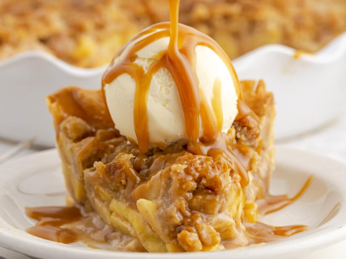 Slice of Caramel Apple Pie topped with a scoop of vanilla ice cream while warm caramel sauce is poured over the cinnamon-spiced apple filling and crumb topping.