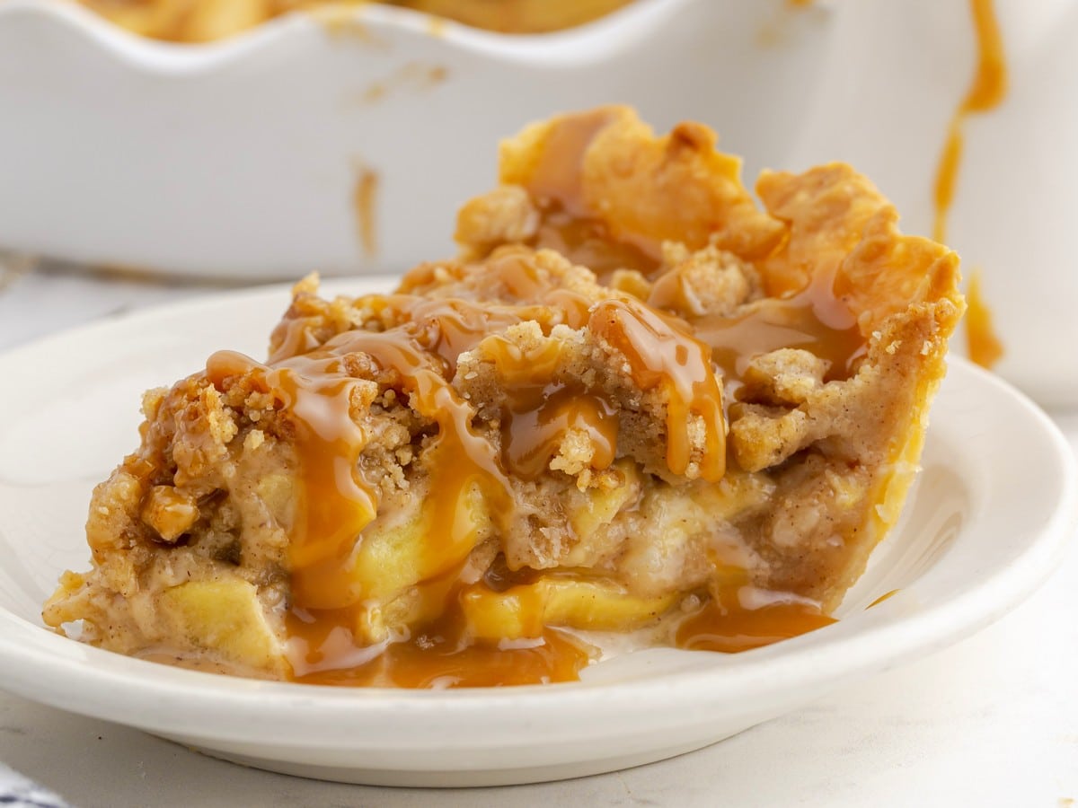 Close-up slice of Caramel Apple Pie on a white plate, showing tender cinnamon-spiced apples beneath a golden crumb topping with caramel sauce generously drizzled over the top and sides.