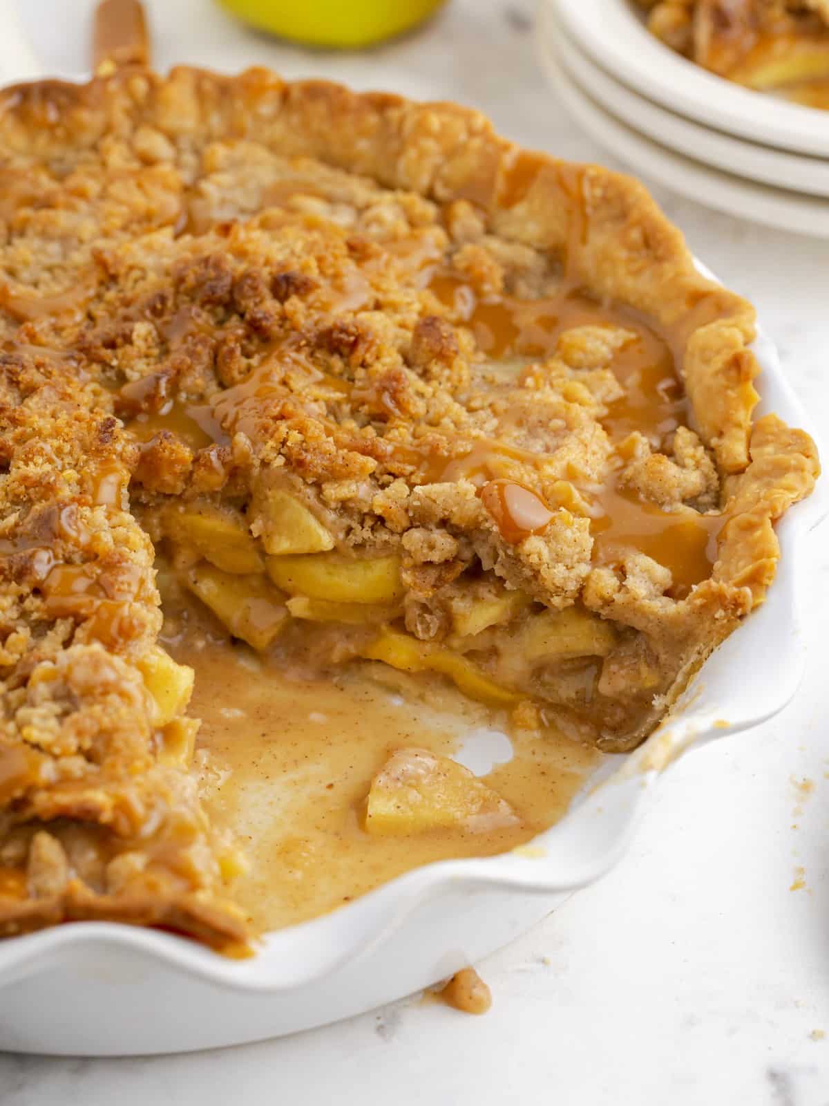 Close-up of Caramel Apple Pie in a white fluted pie dish with a slice removed, revealing soft layered apples, a buttery crumb topping, and caramel sauce pooled over the filling and crust.
