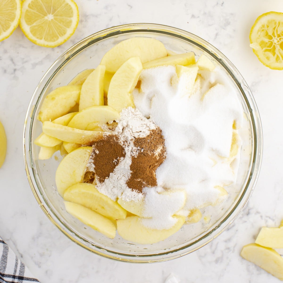 Overhead view of peeled apple slices topped with granulated sugar, flour, cinnamon, and nutmeg in a glass bowl for the Caramel Apple Pie filling.