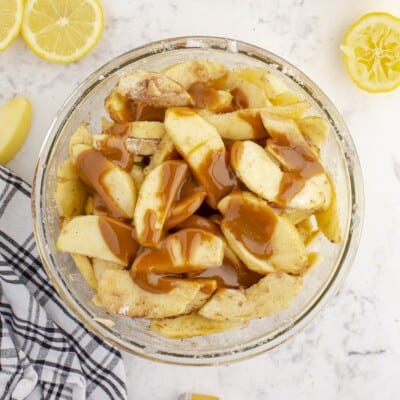 Overhead view of sliced apples coated with cinnamon and sugar and drizzled with caramel sauce in a glass mixing bowl while preparing Caramel Apple Crumb Pie.
