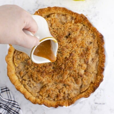 Overhead view of caramel sauce being poured from a small pitcher over a freshly baked Caramel Apple Crumb Pie with a golden brown streusel topping and flaky crust.