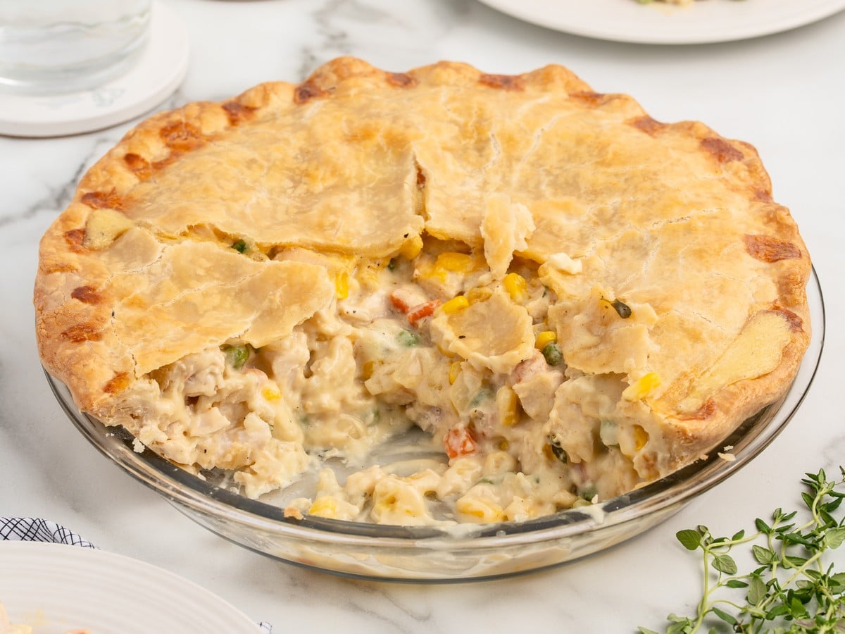 Homemade Chicken Pot Pie in a glass pie dish with a golden, flaky double crust and a creamy filling of chicken, corn, peas, and carrots, with one slice removed.