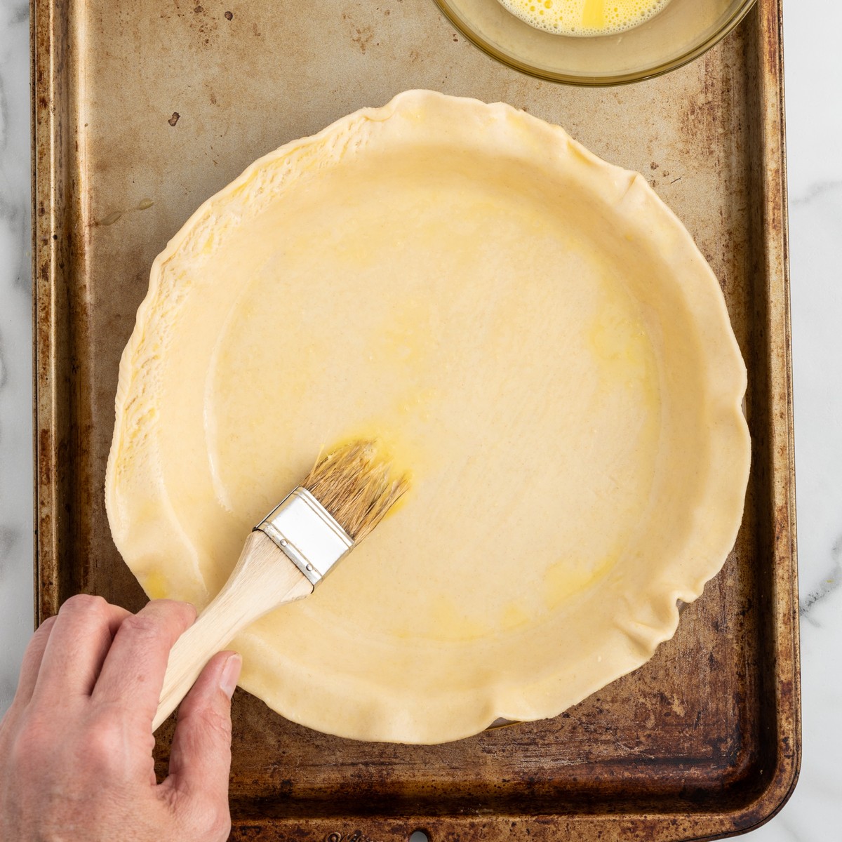 Overhead view of the top pie crust being brushed with egg wash while assembling a homemade Chicken Pot Pie on a baking sheet.