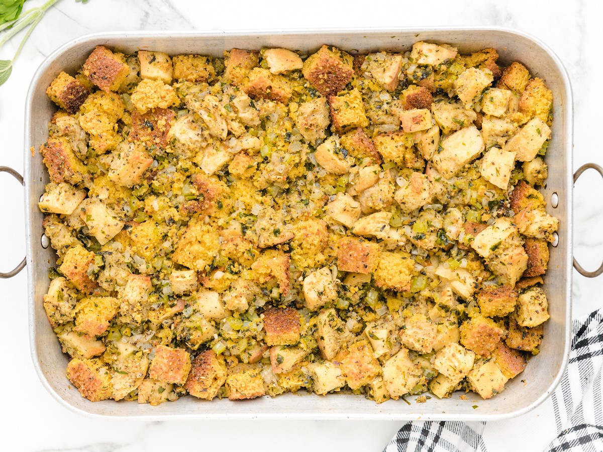 Overhead view of freshly baked Cornbread Stuffing in a large roasting pan with golden cornbread and bread cubes, celery, onion, herbs, and crisp browned edges.