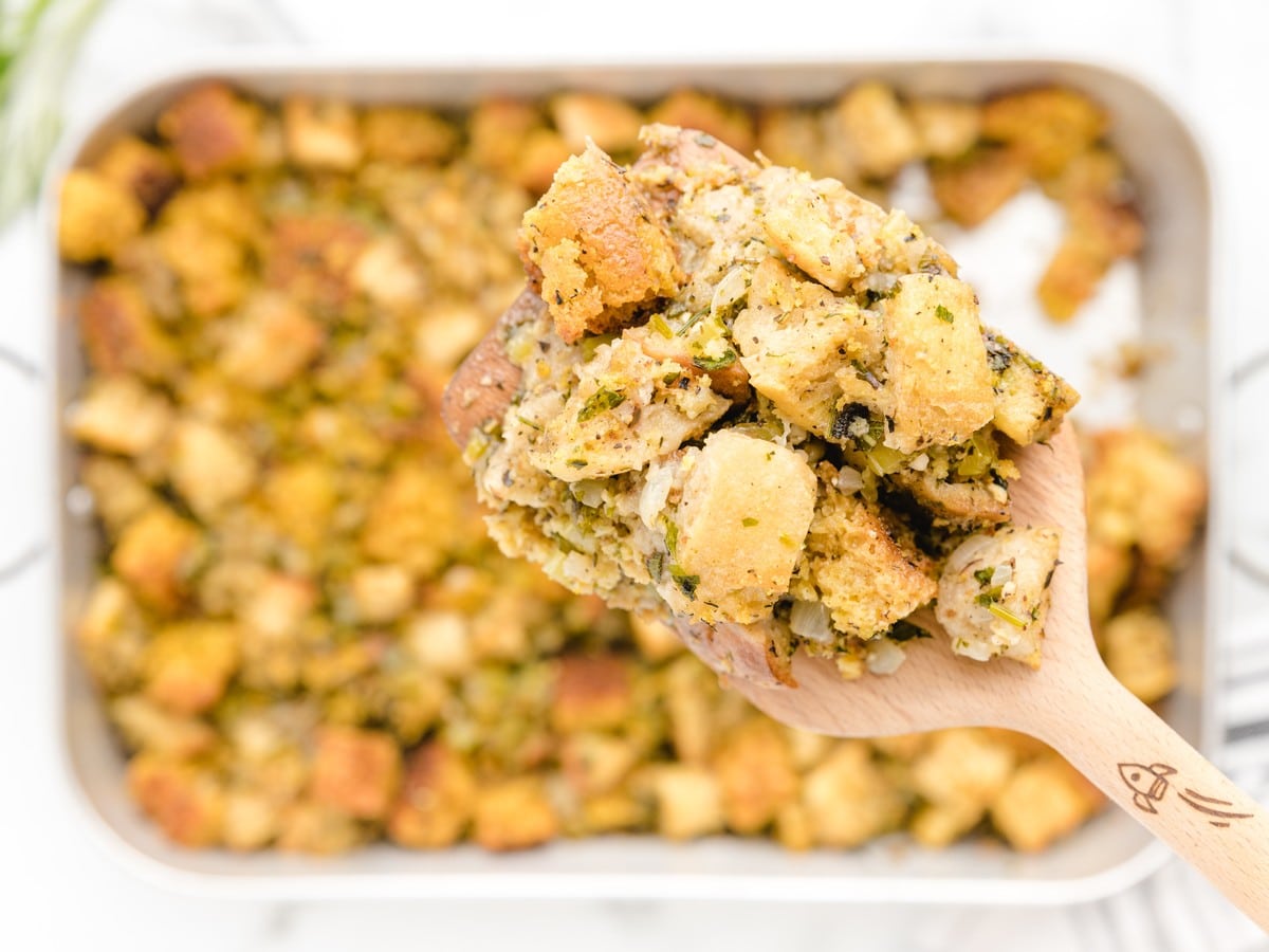 Close-up of a wooden spoon holding a generous serving of Cornbread Stuffing with golden bread and cornbread cubes, celery, onion, and herbs above the baking pan.
