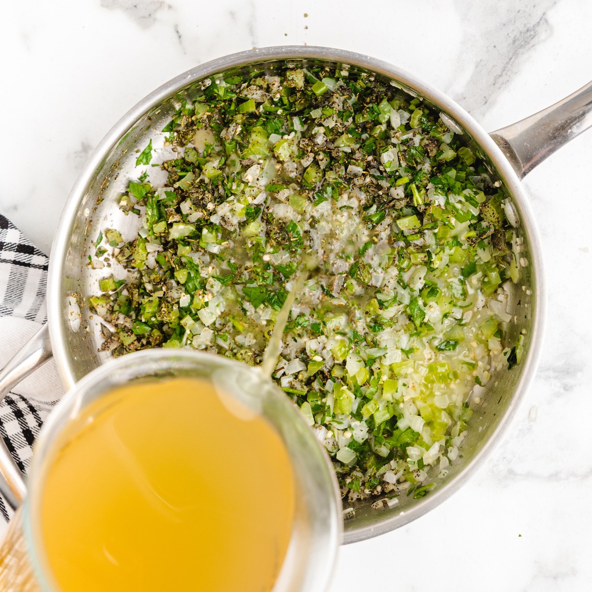 Chicken broth being poured into a skillet of cooked celery, onion, parsley, sage, and seasonings while preparing Cornbread Stuffing.