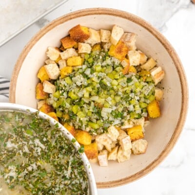 Cornbread and bread cubes topped with the cooked celery, onion, and herb mixture in a large bowl while preparing Cornbread Stuffing.