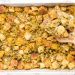 Finished Cornbread Stuffing in a large baking pan with golden cornbread and bread cubes, celery, onion, and herbs, with a wooden spoon holding a serving.