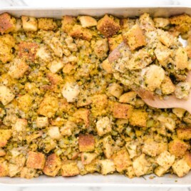 Finished Cornbread Stuffing in a large baking pan with golden cornbread and bread cubes, celery, onion, and herbs, with a wooden spoon holding a serving.