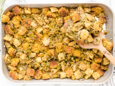 Finished Cornbread Stuffing in a large baking pan with golden cornbread and bread cubes, celery, onion, and herbs, with a wooden spoon holding a serving.
