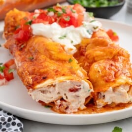Close-up of Cream Cheese Chicken Enchiladas on a white plate with a rich creamy chicken filling, melted cheese, red enchilada sauce, sour cream, fresh diced tomatoes, and chopped cilantro.