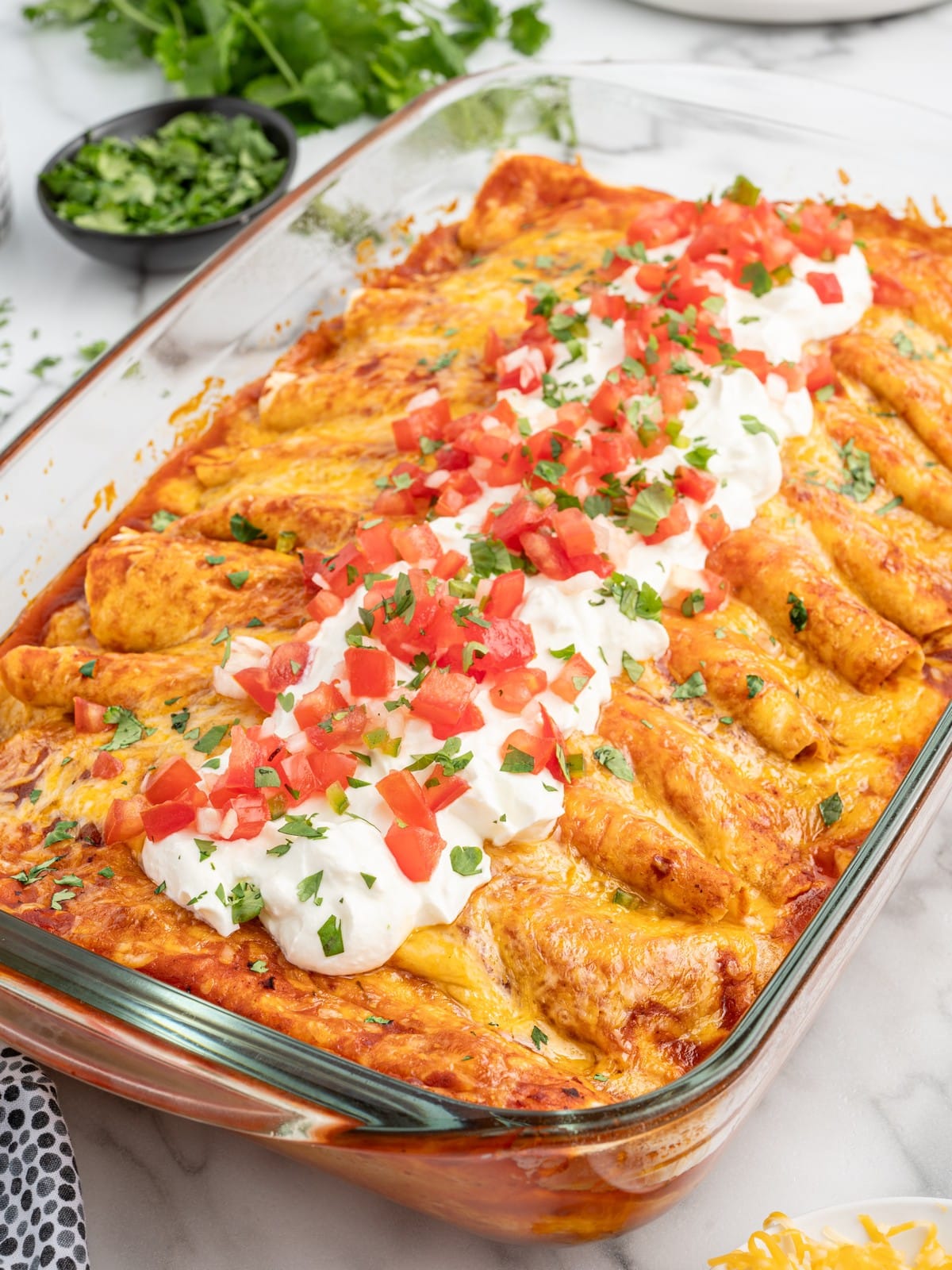 Cream Cheese Chicken Enchiladas baked in a glass casserole dish with melted cheese and red enchilada sauce, topped with sour cream, diced tomatoes, and chopped cilantro.