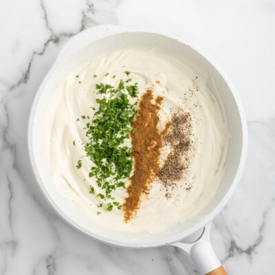 Creamy cream cheese sauce in a skillet topped with chopped cilantro, ground cumin, and black pepper during preparation of Cream Cheese Chicken Enchiladas.