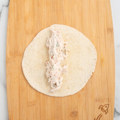 Flour tortilla on a wooden cutting board with a strip of creamy shredded chicken filling placed down the center, ready to be rolled for Cream Cheese Chicken Enchiladas.
