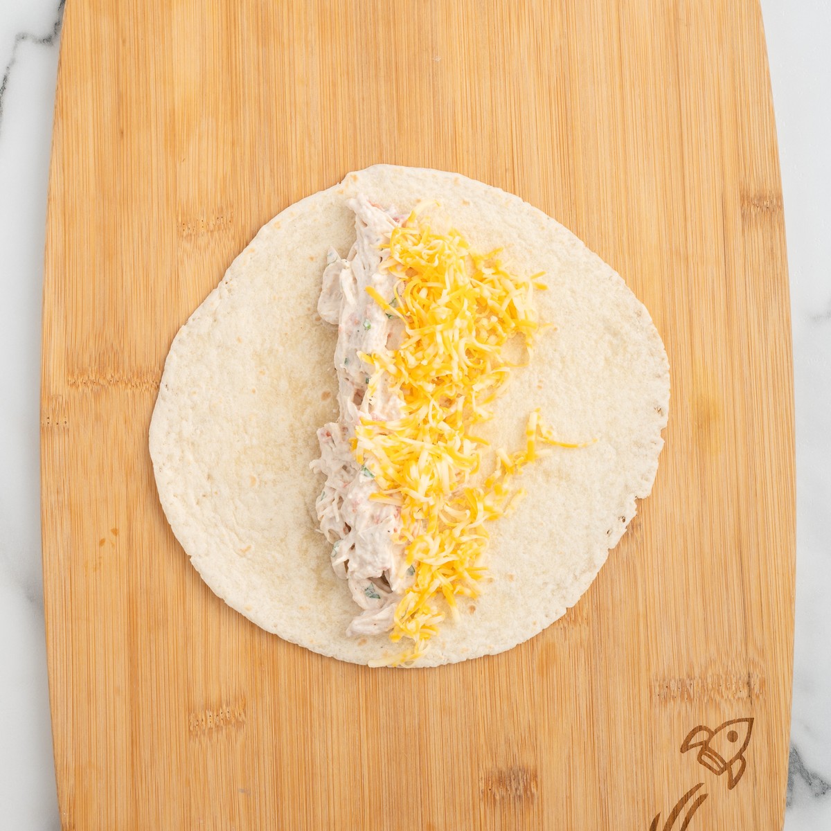 Flour tortilla topped with creamy shredded chicken filling and shredded Colby Jack cheese, ready to be rolled for Cream Cheese Chicken Enchiladas.