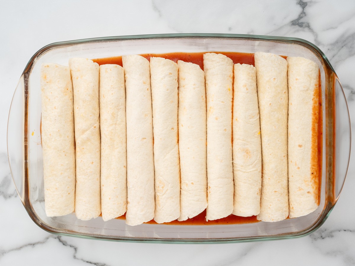 Ten rolled flour tortillas lined up seam-side down in a glass baking dish over a layer of red enchilada sauce for Cream Cheese Chicken Enchiladas.