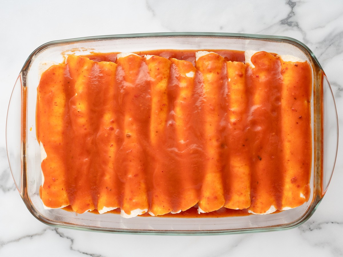 Rolled Cream Cheese Chicken Enchiladas arranged in a glass baking dish and evenly covered with red enchilada sauce before adding cheese and baking.