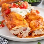 Two Cream Cheese Chicken Enchiladas on a white plate, filled with creamy shredded chicken and topped with melted cheese, red enchilada sauce, sour cream, diced tomatoes, and cilantro.