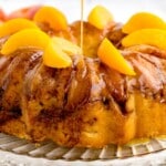 Caramel sauce being drizzled over a peach cobbler pound cake topped with peach slices on a glass cake stand
