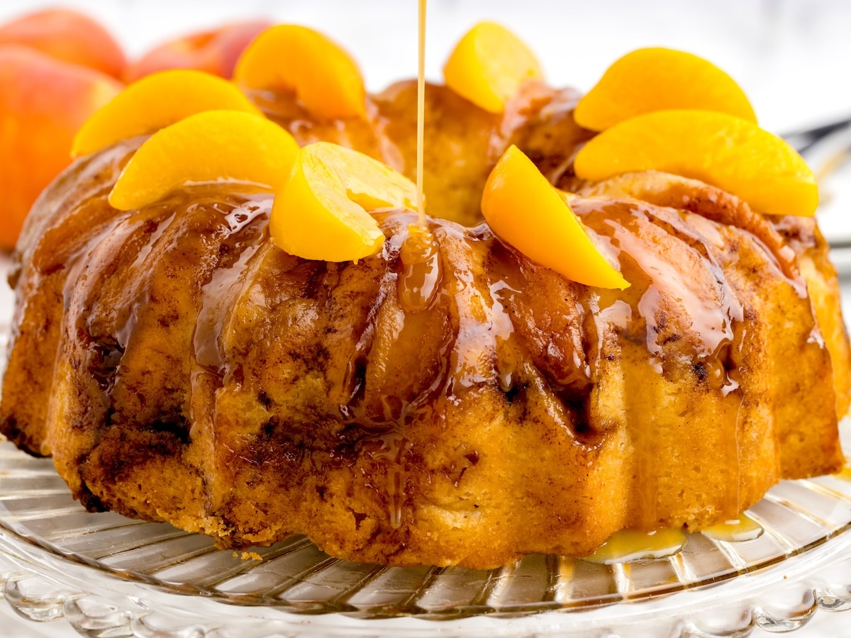 Caramel sauce being drizzled over a peach cobbler pound cake topped with peach slices on a glass cake stand