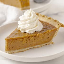 Sweet Potato Pie slice topped with whipped cream, showing a smooth orange-brown filling and flaky crust on a white serving plate.