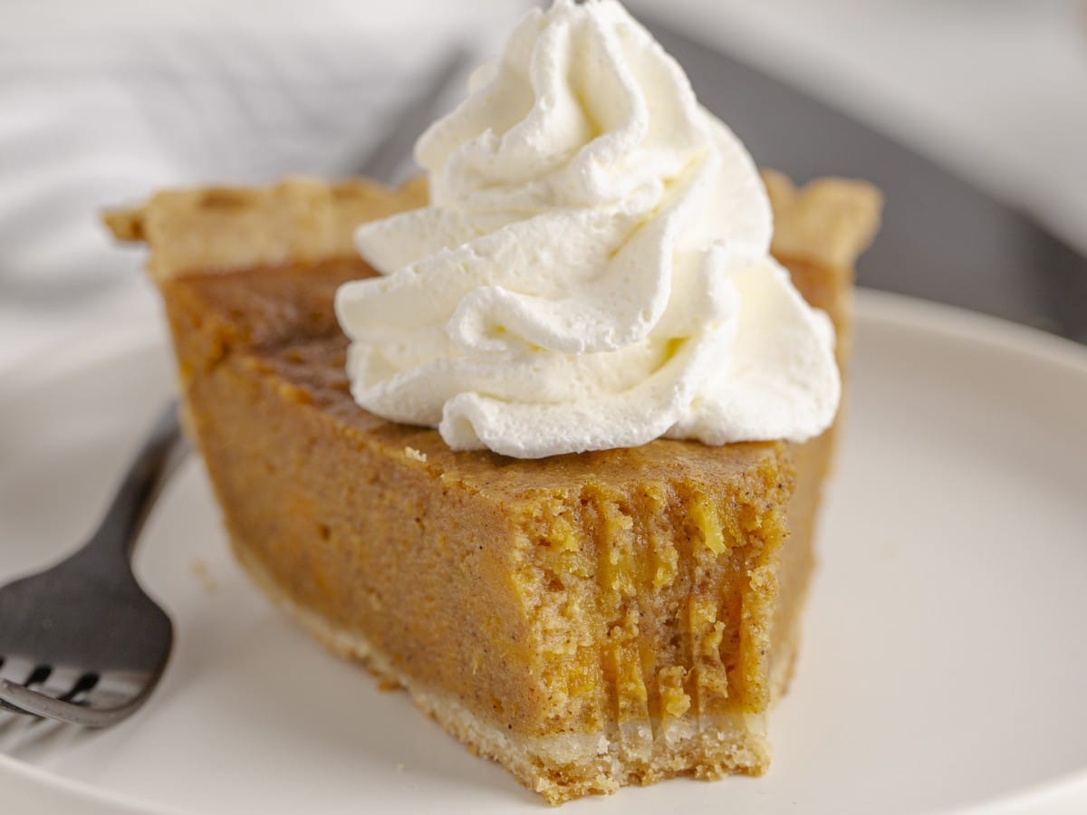 Close-up of a slice of Sweet Potato Pie with smooth spiced filling, a flaky golden crust, and a tall swirl of whipped cream on top.