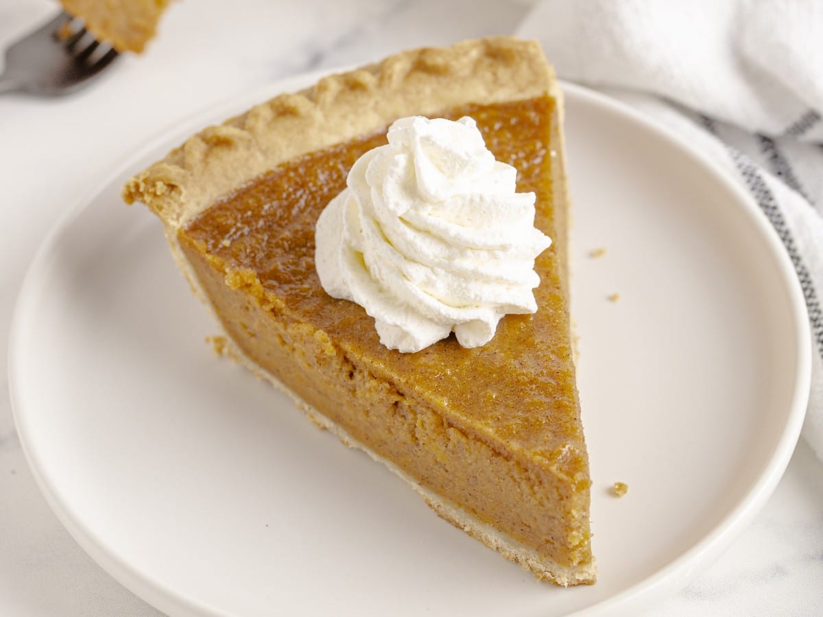 Slice of Sweet Potato Pie served on a white plate with smooth cinnamon-spiced filling, flaky crust, and a swirl of whipped topping.