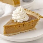 Slice of homemade Sweet Potato Pie with smooth spiced filling, flaky crust, and a swirl of whipped topping on a white plate.