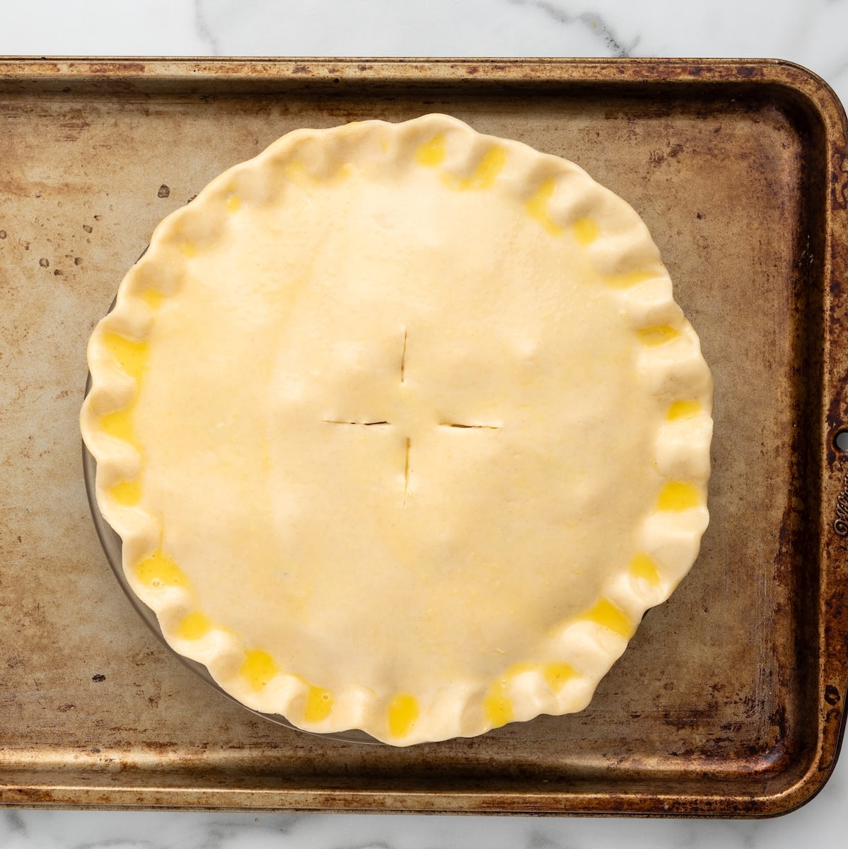 An unbaked chicken pot pie on a baking sheet. The top crust is brushed with egg wash and has small slits in the top for steam to escape.